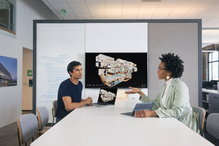 Deux professionnels collaborent dans un bureau moderne pour analyser un projet architectural en 3D. À gauche, un homme consulte un ordinateur portable tandis qu’une femme, à droite, pointe du doigt un écran géant affichant une maquette numérique détaillée d’un bâtiment (vue éclatée des étages et des espaces intérieurs). L’environnement de travail est épuré, avec des éléments graphiques abstraits en arrière-plan (formes géométriques, lignes dynamiques). Idéal pour illustrer les thèmes de la collaboration en équipe, de la visualisation 3D, de l’architecture digitale, ou des outils BIM (Building Information Modeling).